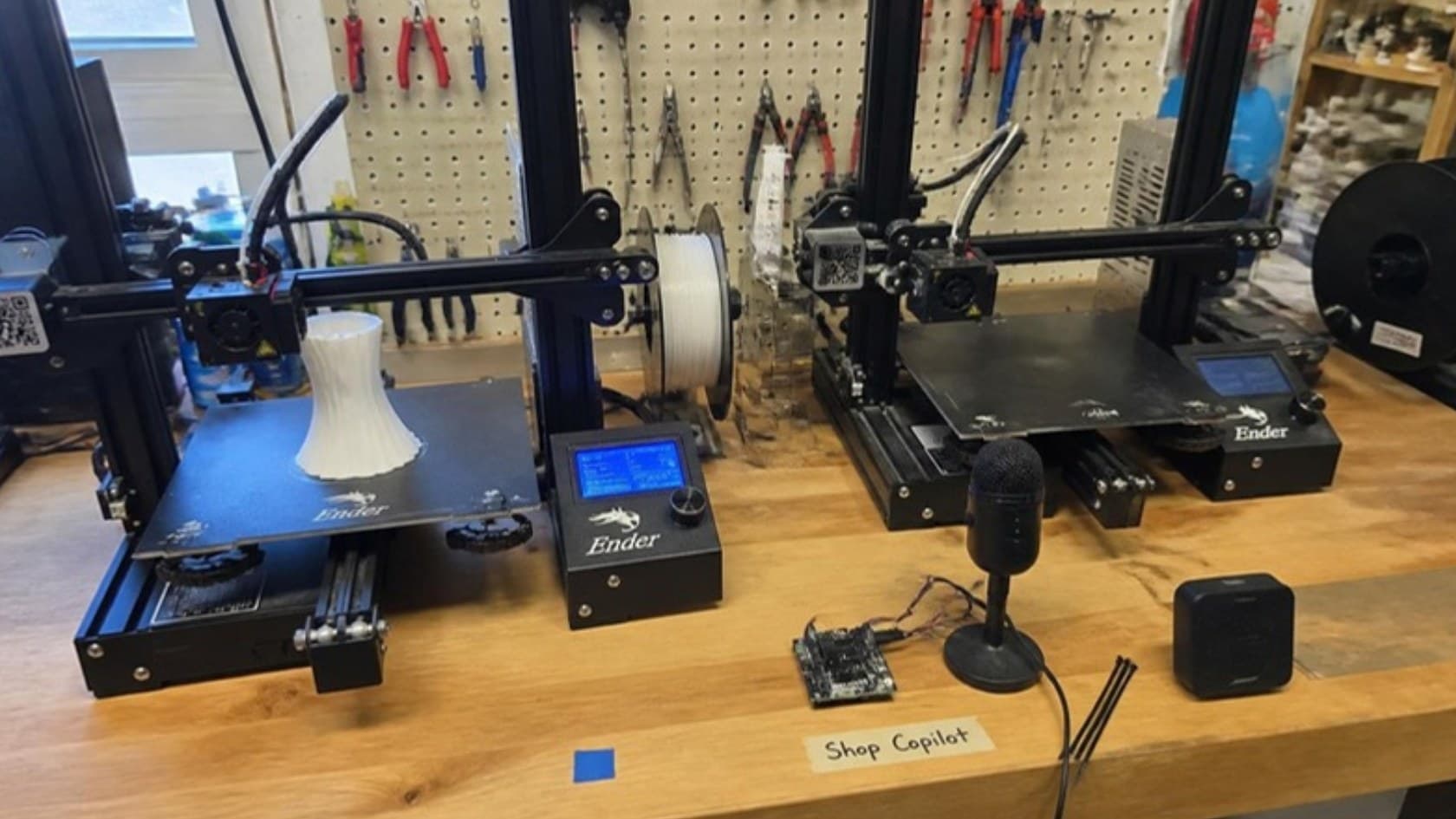 Two Ender 3 printers on a makerspace workbench with a small microphone, speaker and dev board labeled Shop Copilot