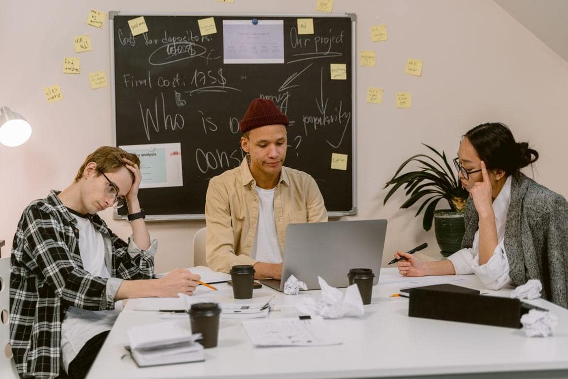 Four students around a table with laptops and notes, talking through a project in front of a chalkboard covered in sticky notes