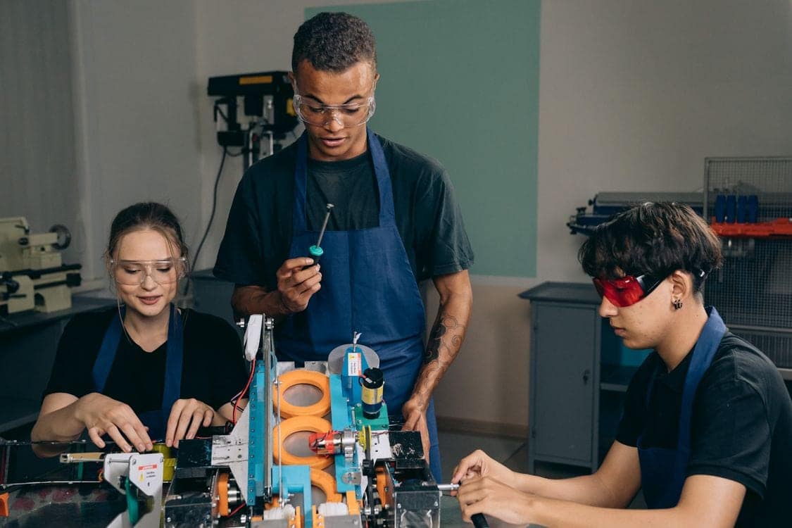 Three students in aprons and safety glasses building a mechanical assembly together at a workshop bench